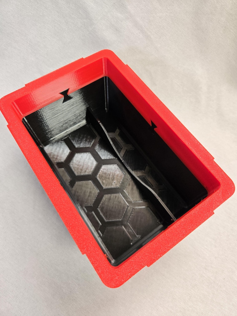 Puede incluir: Un contenedor rectangular rojo y negro con una superficie texturizada. El interior es negro con un patr&oacute;n de panal en la base. El marco rojo tiene detalles recortados en los lados.