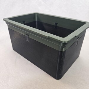 Puede incluir: Un contenedor de almacenamiento rectangular con un cuerpo gris oscuro y un borde gris m&aacute;s claro. El contenedor tiene una superficie texturizada y peque&ntilde;os recortes decorativos en los lados. El interior es oscuro.