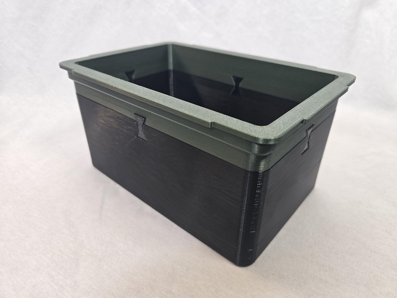 Puede incluir: Un contenedor de almacenamiento rectangular con un cuerpo gris oscuro y un borde gris m&aacute;s claro. El contenedor tiene una superficie texturizada y peque&ntilde;os recortes decorativos en los lados. El interior es oscuro.