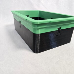 Puede incluir: Contenedor de almacenamiento rectangular de dos tonos. La secci&oacute;n superior es verde, mientras que la inferior es negra. El contenedor tiene detalles recortados y probablemente est&aacute; impreso en 3D.