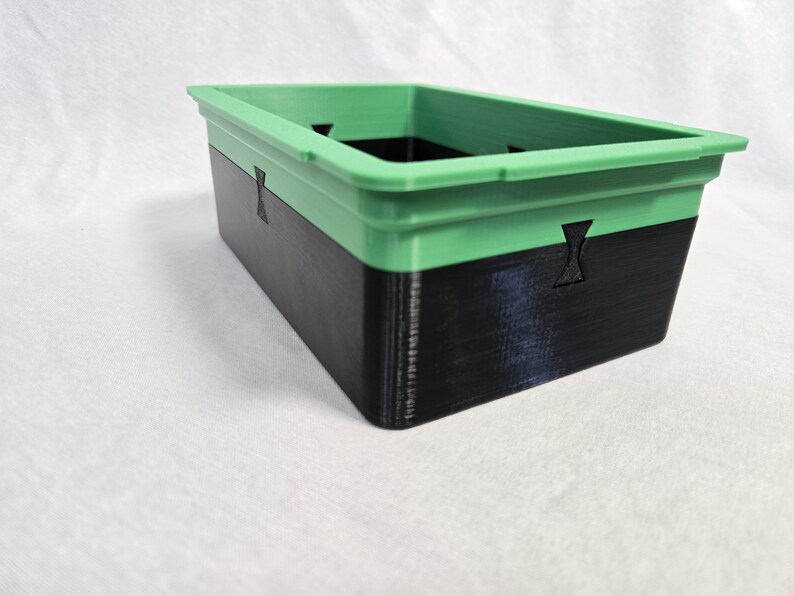 Puede incluir: Contenedor de almacenamiento rectangular de dos tonos. La secci&oacute;n superior es verde, mientras que la inferior es negra. El contenedor tiene detalles recortados y probablemente est&aacute; impreso en 3D.