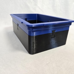 Puede incluir: Contenedor de almacenamiento rectangular de dos tonos. La secci&oacute;n superior es azul vibrante, mientras que la inferior es negra. El contenedor tiene una superficie texturizada y una abertura rectangular. Formas recortadas en los lados.