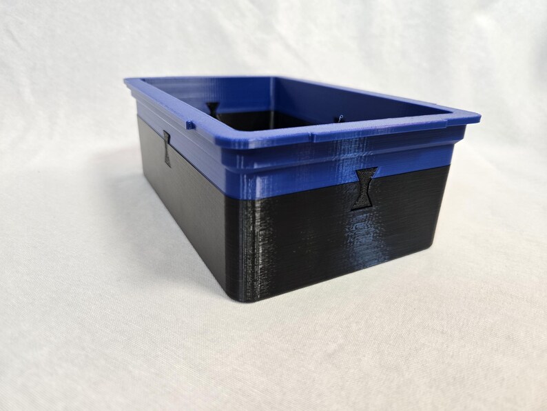 Puede incluir: Contenedor de almacenamiento rectangular de dos tonos. La secci&oacute;n superior es azul vibrante, mientras que la inferior es negra. El contenedor tiene una superficie texturizada y una abertura rectangular. Formas recortadas en los lados.