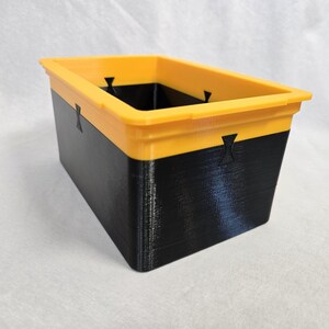 Puede incluir: Contenedor de almacenamiento rectangular de dos tonos. El borde superior es amarillo brillante y la secci&oacute;n inferior es negra. El contenedor tiene una superficie texturizada y probablemente est&aacute; hecho de pl&aacute;stico o un material similar.