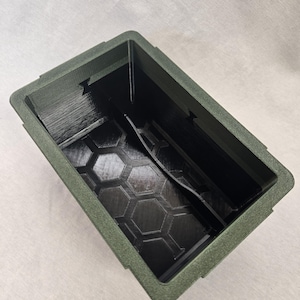 Puede incluir: Un contenedor rectangular verde oliva con un exterior texturizado y un interior negro. El interior presenta un patr&oacute;n de panal en la base y un divisor central. El contenedor est&aacute; vac&iacute;o y visto desde arriba.