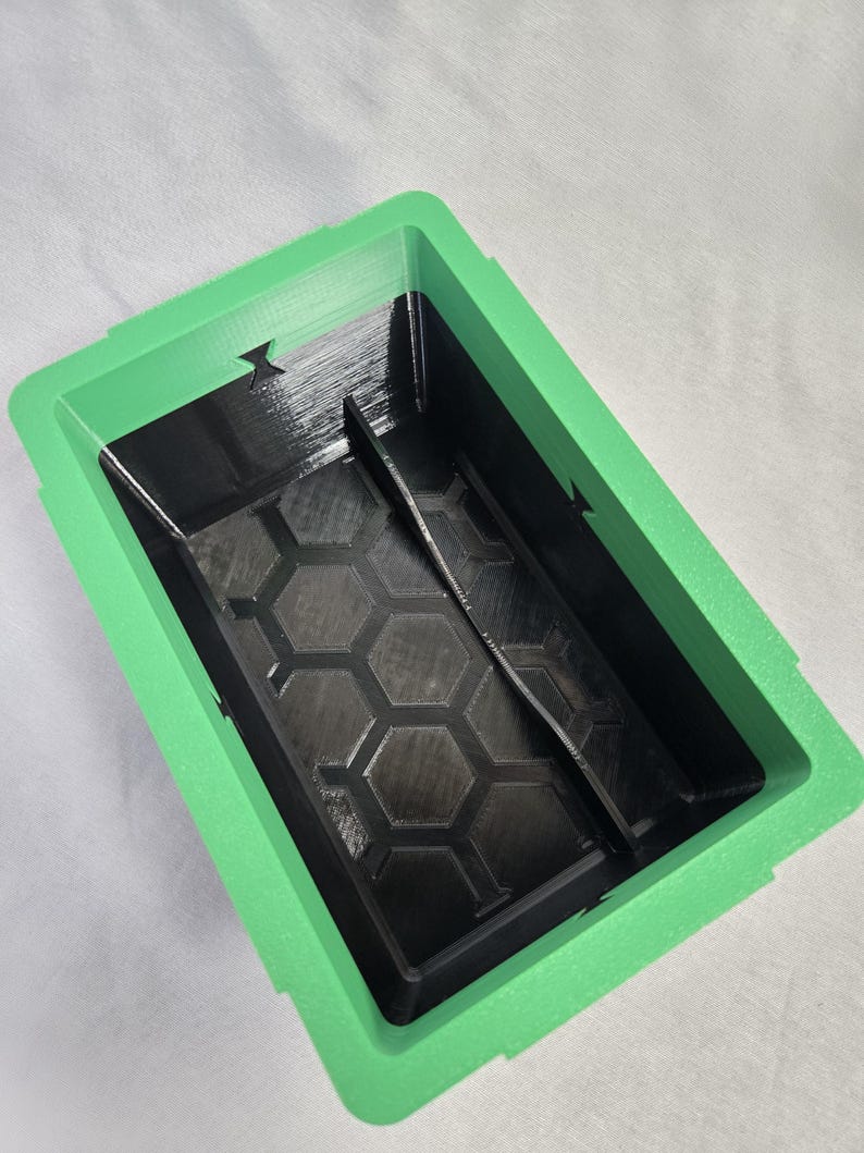 Puede incluir: Un contenedor rectangular de dos tonos con un marco verde y un interior negro. El interior presenta un patr&oacute;n de panal y un divisor central. El contenedor est&aacute; dise&ntilde;ado con una superficie texturizada.