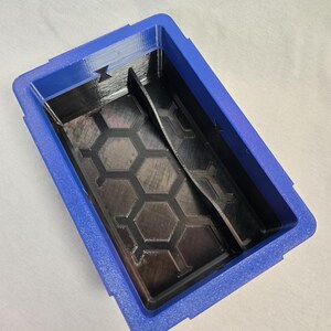 Puede incluir: Un organizador azul y negro impreso en 3D con forma rectangular. El interior presenta un patr&oacute;n de panal y un divisor. El organizador est&aacute; dise&ntilde;ado para guardar objetos peque&ntilde;os y tiene una superficie texturizada.