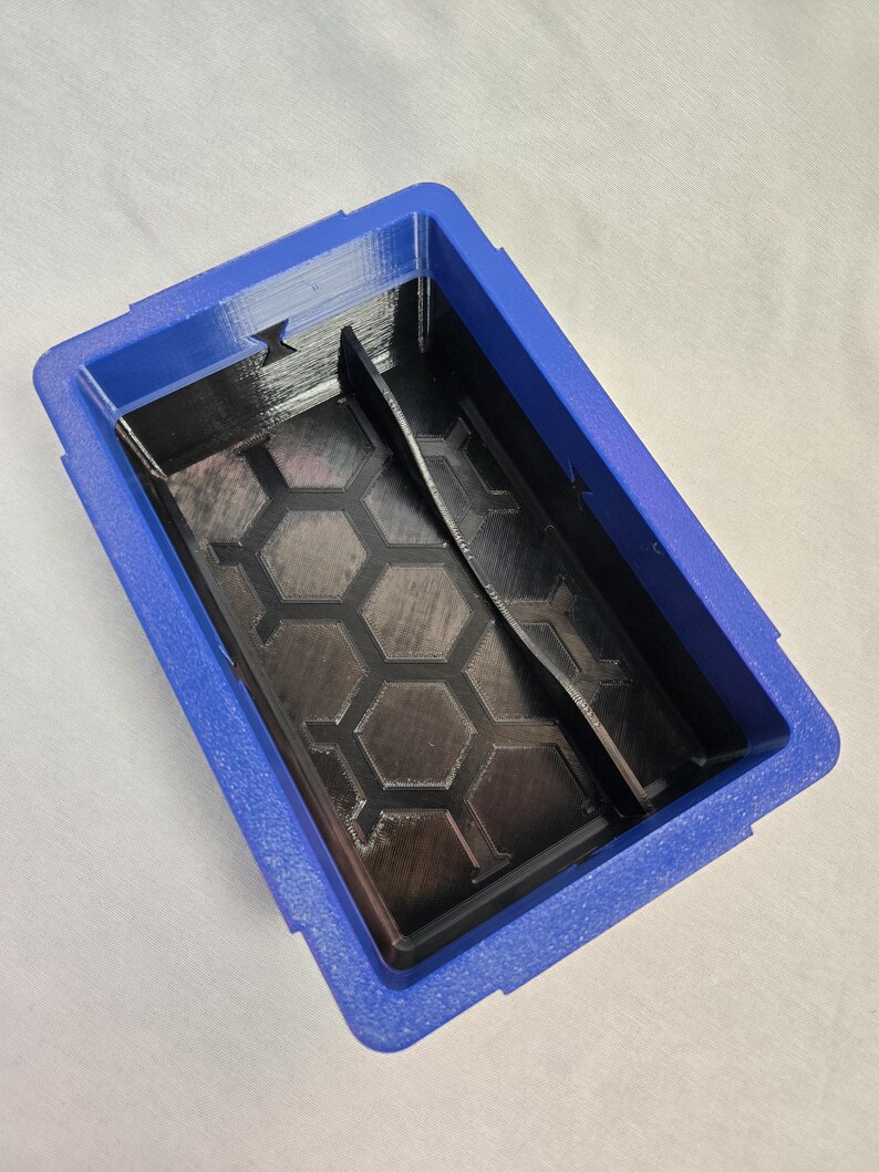 Puede incluir: Un organizador azul y negro impreso en 3D con forma rectangular. El interior presenta un patr&oacute;n de panal y un divisor. El organizador est&aacute; dise&ntilde;ado para guardar objetos peque&ntilde;os y tiene una superficie texturizada.