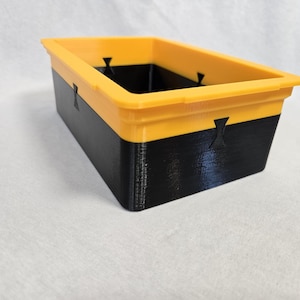 Puede incluir: Contenedor de almacenamiento rectangular de dos tonos. El borde superior es amarillo brillante, mientras que la parte inferior es negra. El contenedor tiene un acabado liso y mate y peque&ntilde;os cortes decorativos en los lados. Dimensiones: 20 cm de largo, 10 cm de ancho, 8 cm de alto.