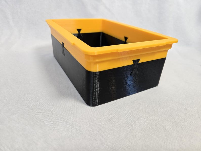 Puede incluir: Contenedor de almacenamiento rectangular de dos tonos. El borde superior es amarillo brillante, mientras que la parte inferior es negra. El contenedor tiene un acabado liso y mate y peque&ntilde;os cortes decorativos en los lados. Dimensiones: 20 cm de largo, 10 cm de ancho, 8 cm de alto.
