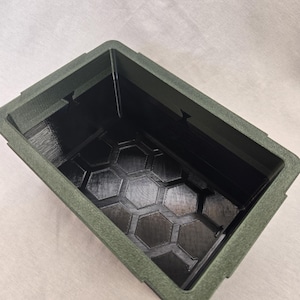 Puede incluir: Un contenedor rectangular verde oscuro con un exterior texturizado y un interior negro. El interior presenta un patrón de panal en la base. El contenedor está vacío y se encuentra sobre una superficie de color claro.