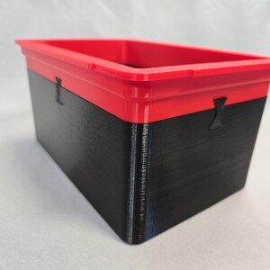 Puede incluir: Un contenedor de almacenamiento rectangular con tapa roja y base negra. El contenedor tiene una superficie texturizada y peque&ntilde;as asas negras en los laterales. El contenedor es probablemente de pl&aacute;stico.