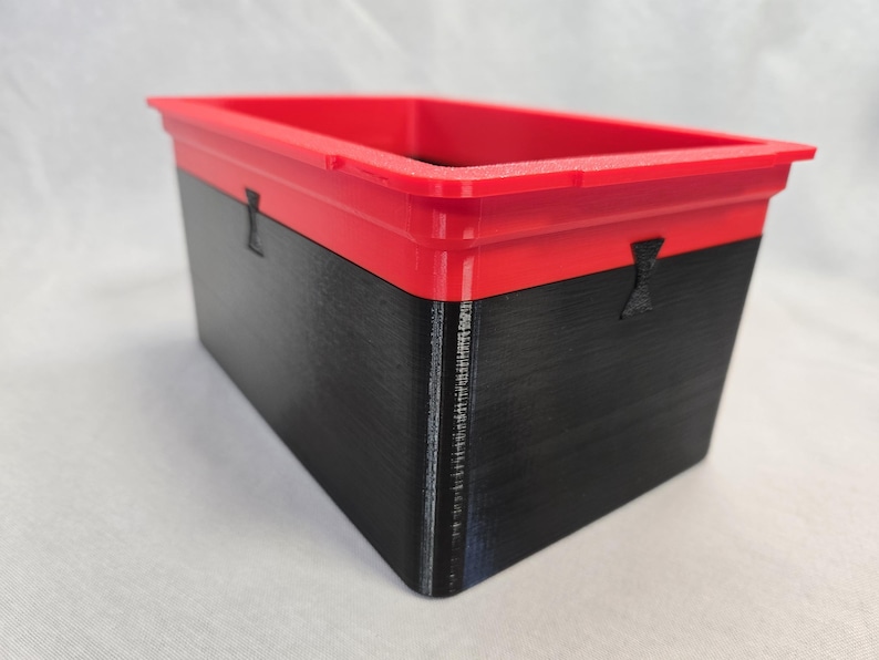 Puede incluir: Un contenedor de almacenamiento rectangular con tapa roja y base negra. El contenedor tiene una superficie texturizada y peque&ntilde;as asas negras en los laterales. El contenedor es probablemente de pl&aacute;stico.