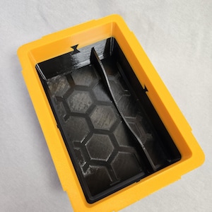 Puede incluir: Un contenedor rectangular de dos tonos, con exterior amarillo e interior negro. El interior presenta un patr&oacute;n de panal y un divisor vertical. El contenedor est&aacute; dise&ntilde;ado con una est&eacute;tica pr&aacute;ctica y utilitaria.