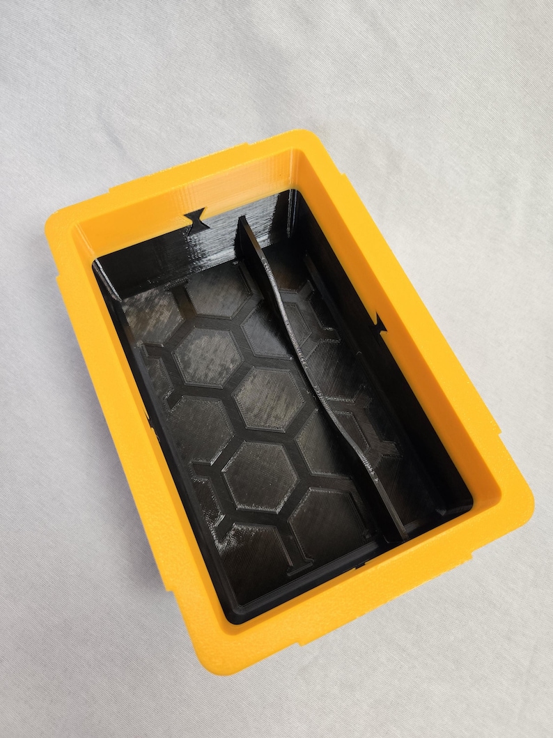 Puede incluir: Un contenedor rectangular de dos tonos, con exterior amarillo e interior negro. El interior presenta un patr&oacute;n de panal y un divisor vertical. El contenedor est&aacute; dise&ntilde;ado con una est&eacute;tica pr&aacute;ctica y utilitaria.