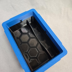 Puede incluir: Un contenedor de almacenamiento rectangular azul y negro. El contenedor tiene un marco exterior azul y un interior negro con un patr&oacute;n de panal. Un divisor negro est&aacute; dentro del contenedor.