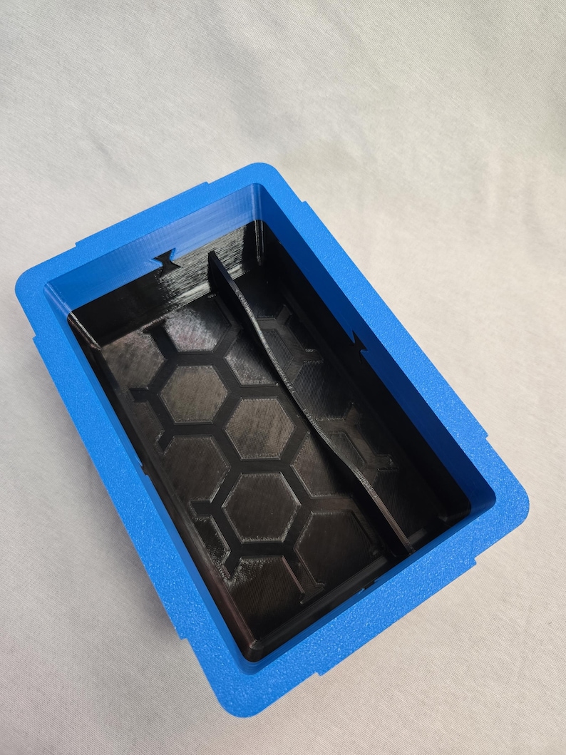 Puede incluir: Un contenedor de almacenamiento rectangular azul y negro. El contenedor tiene un marco exterior azul y un interior negro con un patr&oacute;n de panal. Un divisor negro est&aacute; dentro del contenedor.