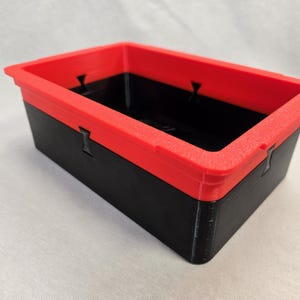 Puede incluir: Un contenedor de almacenamiento rectangular con una base negra y un borde rojo brillante. El contenedor tiene una superficie texturizada y est&aacute; dise&ntilde;ado con elementos entrelazados. El dise&ntilde;o general es simple y funcional, adecuado para organizar objetos peque&ntilde;os.