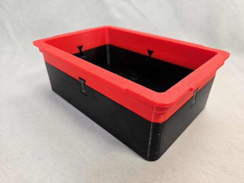 Puede incluir: Un contenedor de almacenamiento rectangular con una base negra y un borde rojo brillante. El contenedor tiene una superficie texturizada y est&aacute; dise&ntilde;ado con elementos entrelazados. El dise&ntilde;o general es simple y funcional, adecuado para organizar objetos peque&ntilde;os.