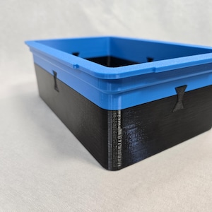 Puede incluir: Un contenedor de almacenamiento rectangular con borde azul y cuerpo negro. El contenedor tiene una superficie texturizada y peque&ntilde;os recortes decorativos. El dise&ntilde;o es moderno y minimalista, adecuado para organizar art&iacute;culos.