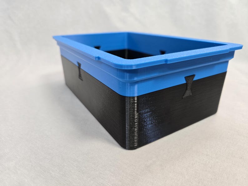 Puede incluir: Un contenedor de almacenamiento rectangular con borde azul y cuerpo negro. El contenedor tiene una superficie texturizada y peque&ntilde;os recortes decorativos. El dise&ntilde;o es moderno y minimalista, adecuado para organizar art&iacute;culos.