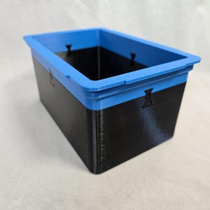 Puede incluir: Un contenedor de almacenamiento rectangular con un cuerpo negro y un borde azul brillante. El contenedor tiene una superficie texturizada y peque&ntilde;os recortes decorativos en los lados. El contenedor est&aacute; dise&ntilde;ado para organizar y almacenar art&iacute;culos.
