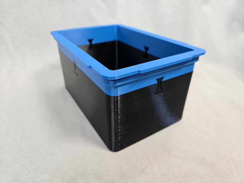 Puede incluir: Un contenedor de almacenamiento rectangular con un cuerpo negro y un borde azul brillante. El contenedor tiene una superficie texturizada y peque&ntilde;os recortes decorativos en los lados. El contenedor est&aacute; dise&ntilde;ado para organizar y almacenar art&iacute;culos.