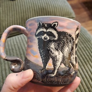 Puede incluir: Una taza de cerámica hecha a mano con una ilustración de mapache. La taza presenta un diseño de mapache en blanco y negro sobre un fondo rosa y azul. El asa tiene un diseño en espiral con detalles en naranja y gris. La taza es sostenida por una persona.