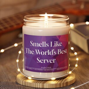 May include: A lit soy candle in a glass jar with a purple label that reads "Smells Like The World's Best Server." The candle sits on a round wooden coaster. A guitar and book are in the background.