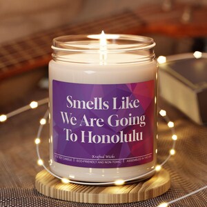 May include: A lit soy candle in a glass jar with a purple label that reads "Smells Like We Are Going To Honolulu." The candle sits on a wooden coaster, with string lights and a guitar in the background.