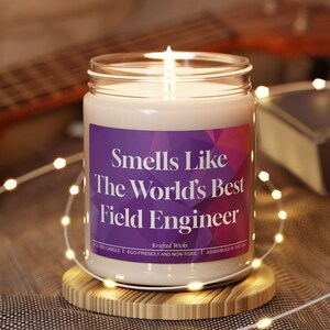 Może przedstawiać: Zapalone świeca sojowa w szklanym słoiku z fioletową etykietą z napisem "Smells Like The World's Best Field Engineer". Świeca stoi na drewnianej podstawce, z łańcuchem świetlnym i gitarą w tle.