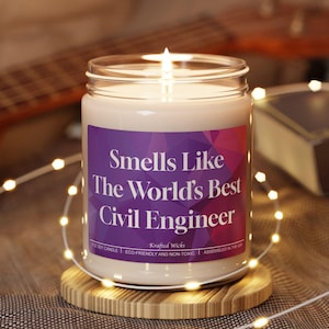 May include: A lit candle in a clear glass jar with a purple label. The label reads "Smells Like The World's Best Civil Engineer." The candle sits on a round wooden coaster. A guitar and a book are in the background.