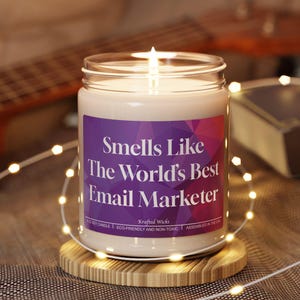 May include: A lit candle in a glass jar with a purple label that reads "Smells Like The World's Best Email Marketer." The candle sits on a round wooden coaster. A guitar and a book are in the background.