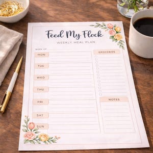 May include: A weekly meal plan titled "Feed My Flock" with floral accents. The planner includes sections for each day of the week, groceries, and notes. A pen, a bowl of pasta, a cup of coffee, and flowers are on the wooden surface.