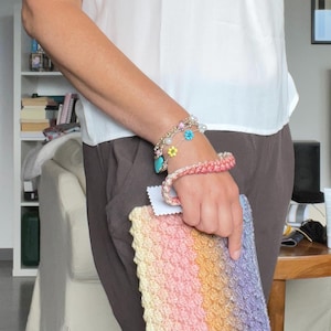 Può includere: Una pochette rettangolare all'uncinetto caratterizzata da un motivo testurizzato a gradiente diagonale nei toni del giallo pallido, rosa tenue, arancione e viola chiaro. La borsa include un cinturino da polso intrecciato e un'etichetta bianca.