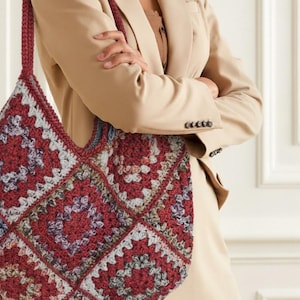 Può includere: Grande borsa tote all'uncinetto fatta a mano con motivo a granny square. La borsa presenta una palette di colori bordeaux, grigio e crema. Il cinturino è in tinta bordeaux. La borsa è tenuta da una persona con un blazer beige.
