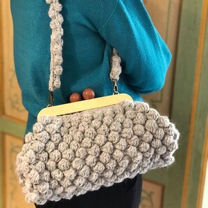 Può includere: Borsa a mano fatta a mano di colore grigio chiaro, con una texture a rilievo. Presenta una chiusura in legno chiaro e una tracolla abbinata. La borsa è portata a spalla.
