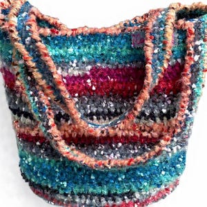 Può includere: Una borsa tote all'uncinetto con strisce orizzontali in tonalità vivaci di verde acqua, magenta, arancione, nero e grigio. La borsa è realizzata in filato testurizzato multicolore dall'aspetto soffice e include due tracolle integrate.