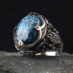 May include: A silver ring with an oval, blue gemstone. The ring has intricate, dark metalwork with gold accents and small turquoise stones around the gemstone. The ring's design is ornate and detailed.