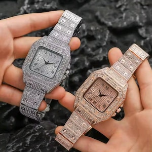 May include: Two luxury watches, one silver and one rose gold, are displayed. Both watches have square faces with Roman numeral hour markers and are encrusted with sparkling crystals. The watch bands are also fully crystal-covered.
