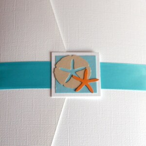 May include: A white card with a turquoise ribbon and a square cutout with a light blue background. Inside the cutout is a tan sand dollar with a white starfish and an orange starfish.