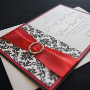 May include: A wedding invitation with a black and white damask pattern, a red satin ribbon, and a rhinestone embellishment. The invitation reads "Emily Marie & Samuel Walt".