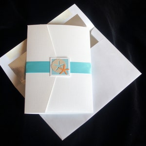 May include: White wedding invitation with a blue ribbon and a cutout of a sand dollar and starfish. The invitation is folded in half and has a white envelope.