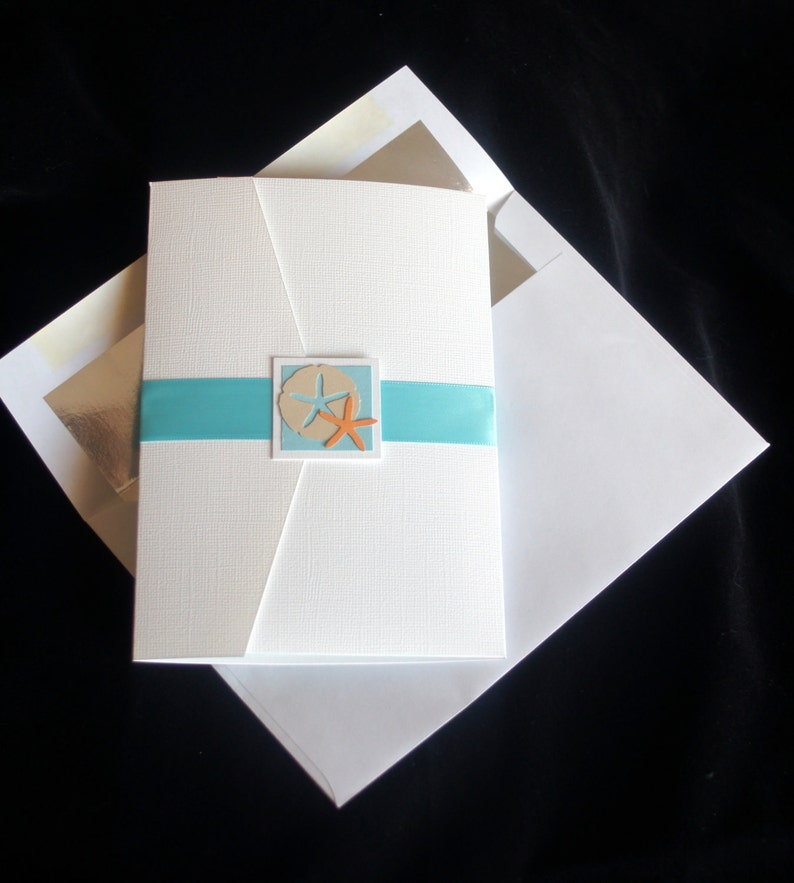 May include: White wedding invitation with a blue ribbon and a cutout of a sand dollar and starfish. The invitation is folded in half and has a white envelope.