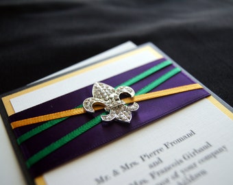May include: A white wedding invitation with a purple, green, and gold ribbon and a silver fleur-de-lis embellishment. The text on the invitation reads "Mr. & Mrs. Pierre Fromand and Ms. Francis Garland".