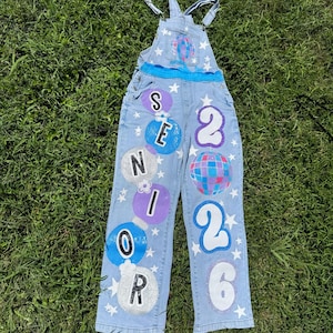 Puede incluir: Overol de mezclilla azul claro decorado con el texto "SENIOR 2026" en blanco y diseños de burbujas coloridas. El overol tiene tirantes ajustables y está adornado con estrellas blancas.
