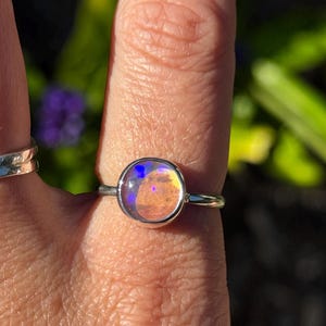 May include: A silver ring featuring a round, iridescent gemstone. The gemstone shows a blend of colours, including blue, orange, and yellow. The ring has a simple, elegant design with a thin band.