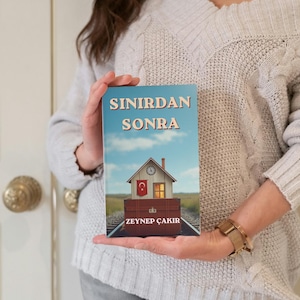 Puede incluir: Portada de libro con el título "SINIRDAN SONRA" en tipografía vintage. La ilustración muestra una casa con una bandera turca sobre una vía de tren, con un cielo azul de fondo. También se muestra el nombre de la autora, ZEYNEP ÇAKIR.