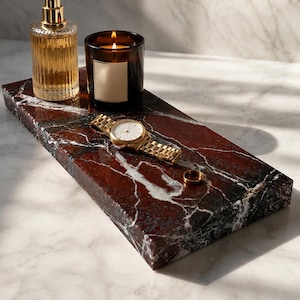 Peut inclure: Un plateau rectangulaire en marbre rouge foncé avec des veines blanches, supportant un flacon de parfum en verre transparent avec un bouchon doré, une bougie allumée dans un pot en verre brun, une montre-bracelet dorée et une bague dorée.