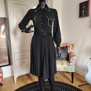 May include: A black long-sleeved dress featuring a pointed collar, button-down front, and two chest pockets with light green piping. The dress includes a matching black belt with a buckle at the waist and a pleated skirt that falls below the knee.